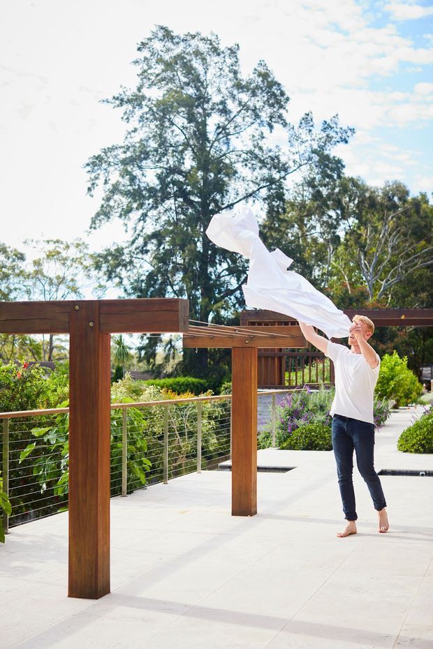 A bright outdoor courtyard or terrace with large timber pergola posts and beams, stainless-steel cable railings, pale paved flooring, and landscaped garden beds. A barefoot person is lifting a flowing white fabric beneath the pergola. Mature trees and a blue sky form the background.
