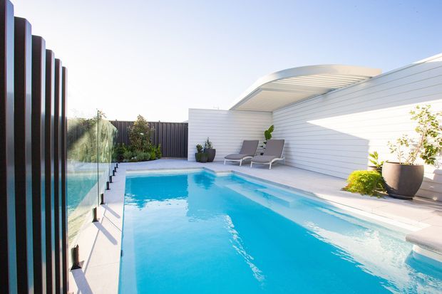 Bright contemporary outdoor pool area with a long rectangular swimming pool, pale paved surrounds, white horizontal wall cladding, a curved translucent roof canopy, dark vertical screening, glass pool fencing, lounge chairs, and potted planting.