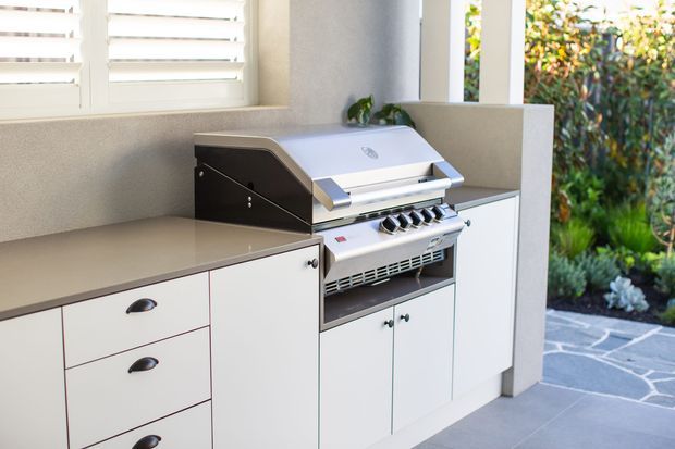 A built-in outdoor kitchen with a stainless-steel barbecue grill set into white cabinetry, drawers, and a neutral stone-look benchtop. The compact cooking area is sheltered by walls and positioned beside a garden, with grey stone paving underfoot and a bright, clean contemporary finish.