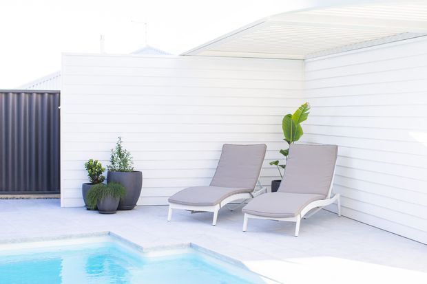 Bright, minimalist outdoor poolside setting with two cushioned reclining loungers arranged beside a pale blue swimming pool. White horizontal cladding and a white paved terrace create a clean, contemporary backdrop, with dark planters and green foliage providing contrast.