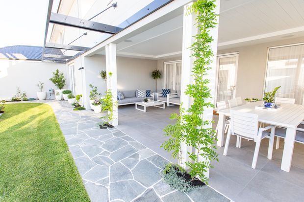 Bright, spacious covered outdoor entertaining area adjoining a contemporary home. The patio features a large white dining setting, a separate outdoor lounge with patterned cushions, pale grey tiled flooring, white structural columns, overhead beams and translucent roof panels. A paved path borders a manicured lawn, with climbing greenery and potted plants adding a fresh garden character.