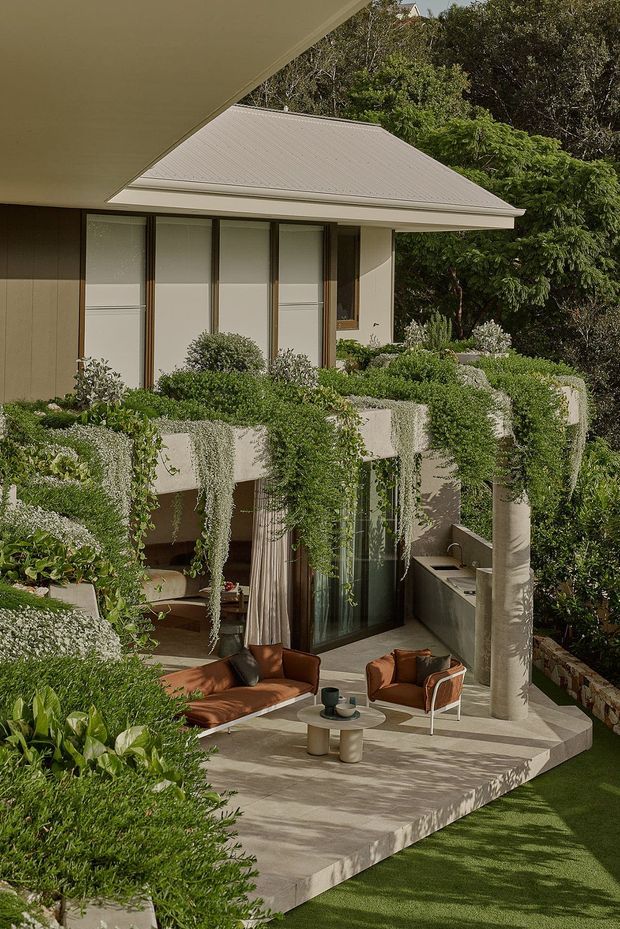A multi-level contemporary home surrounded by dense landscaped greenery. The image focuses on an outdoor terrace with a concrete-paved sitting area, curved concrete columns, lush cascading plantings, a glazed lower-level facade, and warm brown outdoor lounge furniture. A light metal hipped roof and vertically framed upper-level glazing are visible above.