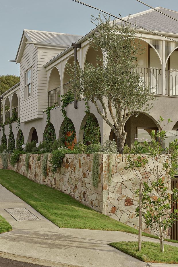 Exterior view of a substantial multi-level residential building with pale horizontal weatherboard cladding, light corrugated metal pitched roofs, repeated arched verandah openings, slender decorative columns, and metal balustrades. A textured natural-stone retaining wall, landscaped garden, mature olive tree, lawn, and concrete footpath occupy the foreground.