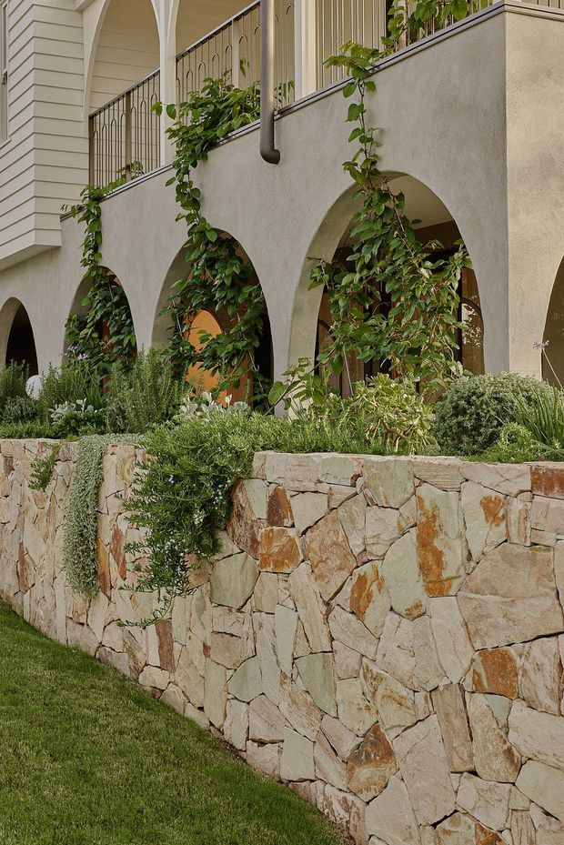 Exterior view of a refined, light-coloured residence with a raised terrace or balcony featuring repeated stucco arches, dark metal railings, climbing vines, and a substantial natural stone retaining wall. Layered drought-tolerant landscaping and a neatly maintained lawn occupy the foreground.