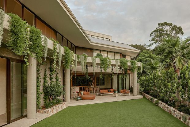 Exterior view of a sophisticated multi-level residence arranged around a landscaped courtyard. The building features broad overhanging roof planes, extensive floor-to-ceiling glazing, warm timber-framed windows and doors, pale cylindrical columns, stone-clad low walls, and a covered outdoor living area furnished with seating. Dense climbing and hanging greenery integrates the architecture with the garden, while a manicured lawn occupies the foreground.