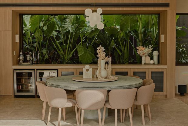 Warm, refined dining room with a large round stone dining table surrounded by upholstered beige dining chairs. Full-height timber-framed glazing looks onto dense tropical foliage, while built-in timber cabinetry and shelving provide storage and display space. A sculptural clustered pendant light hangs above the table, and neutral stone, timber, and cream furnishings create a sophisticated indoor-outdoor atmosphere.