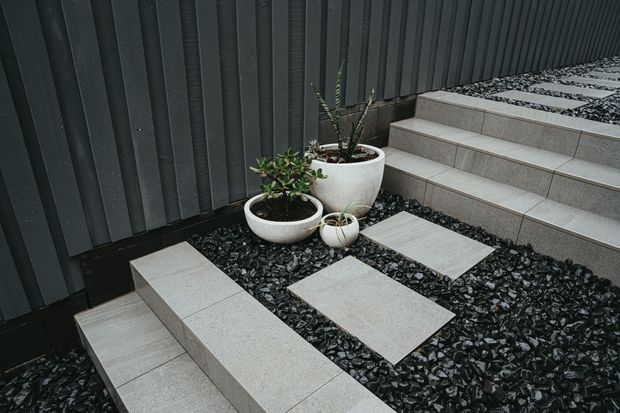 A contemporary exterior courtyard or entry landscape featuring dark vertical metal cladding, pale tiled steps and raised rectangular stepping platforms set in a bed of dark decorative stone. White rounded planters with succulents provide a subtle natural accent.