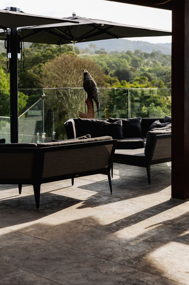 A covered outdoor lounge or terrace with dark upholstered seating arranged around a low table, a glass balustrade, large umbrellas, and a timber post. The space has a paved stone or concrete floor and overlooks a lush wooded landscape. A bird sculpture or carving stands near the balustrade.
