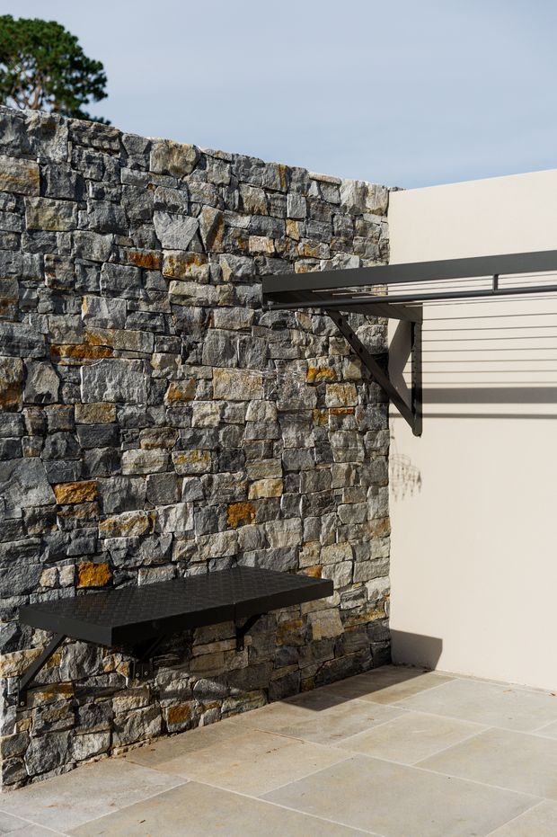 Exterior architectural detail showing a rugged stacked-stone masonry wall beside a smooth off-white rendered wall. A black metal cantilevered bench or shelf and a slim metal awning structure project from the wall, with large pale stone paving underfoot. The composition is minimalist and contemporary, with strong geometric lines and contrasting rough natural stone against smooth plaster.