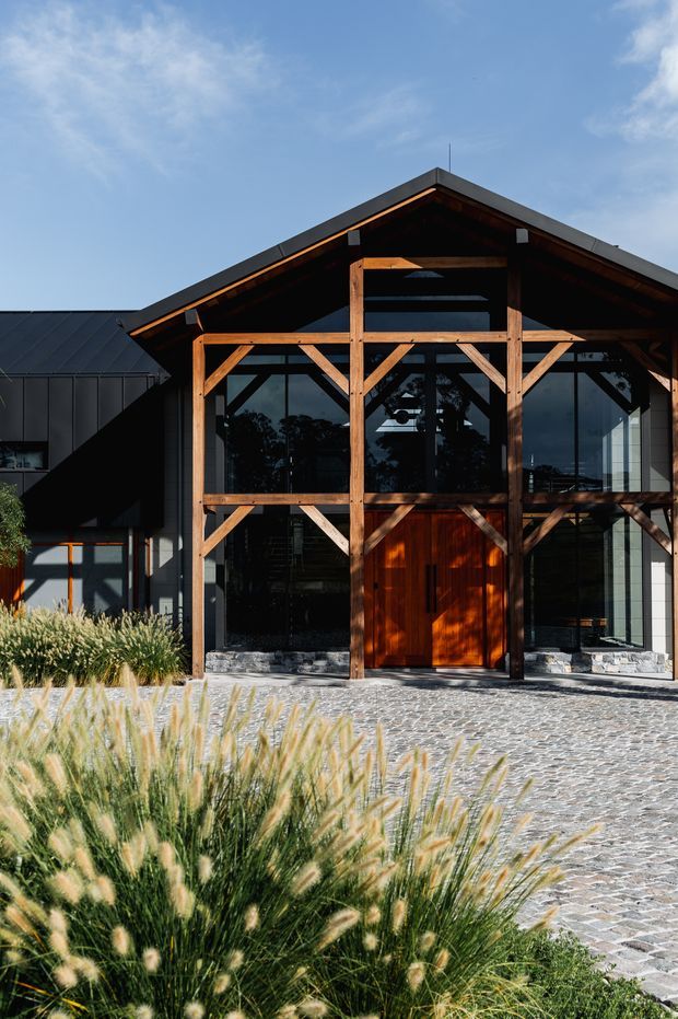 Exterior view of a contemporary gabled building with a prominent exposed timber post-and-beam facade, extensive dark-framed glazing, and a warm timber entry door. The structure is set behind a broad cobblestone forecourt with ornamental grasses in the foreground. The building combines dark metal cladding with a timber-framed entrance and pitched roof.