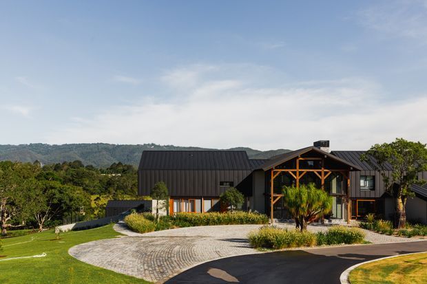 A contemporary dark-clad residential house set in a landscaped suburban or semi-rural site. The building has multiple steep pitched gable roofs, prominent timber-framed glazing at the entry, and a broad curved cobblestone driveway with lawn and ornamental planting. Tree-covered hills are visible in the background.