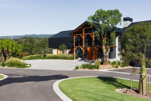 Exterior view of a contemporary dark-clad house with a prominent timber post-and-beam entry structure, steeply pitched roof elements, and landscaped grounds. A pale paved driveway and curved asphalt access road lead to the entrance, with mature and newly planted trees surrounding the property.