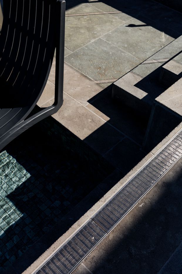 Close-up architectural detail of a dark tiled pool edge or sunken water feature beside a paved terrace. A dark outdoor lounge chair is partially visible at left, with strong sunlight casting geometric shadows across the stone paving and linear drainage grate.