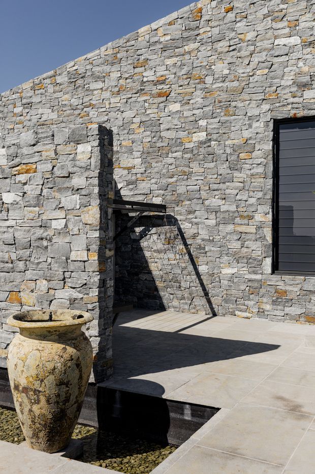 A sunlit exterior courtyard or terrace enclosed by a rustic dry-stacked grey stone wall. Large pale stone paving surrounds a recessed dark water feature or channel filled with pebbles, while a weathered stone planter stands in the foreground. A black-framed horizontal-panel door or window is set into the wall, and a simple metal awning casts strong geometric shadows across the paving.