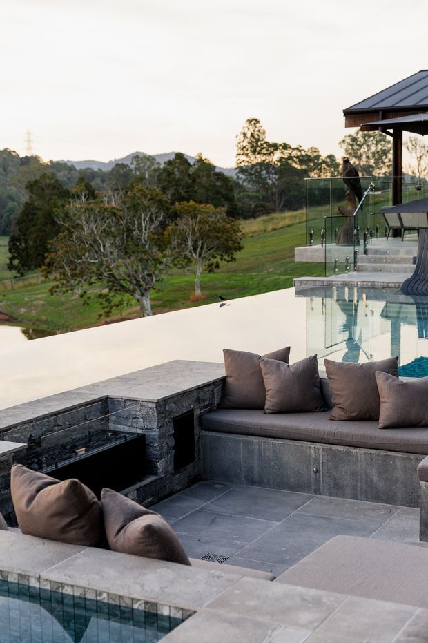 A sophisticated outdoor entertaining area overlooking a landscaped rural hillside. The terrace features a swimming pool, dark stone paving and built-in stone seating with charcoal upholstered cushions, a sunken outdoor lounge, a linear fire feature, glass balustrades, and a covered pavilion with a dark metal roof. The setting is serene and contemporary, with expansive views across greenery and distant hills.