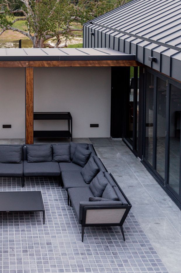 A modern courtyard terrace viewed from above, featuring a dark grey L-shaped outdoor sectional sofa around a low rectangular coffee table. The seating area sits on small square grey pavers, with larger grey floor tiles extending toward full-height dark-framed glazing. White rendered walls, warm timber posts and beams, and a projecting roof clad in dark grey standing-seam metal create a refined contemporary outdoor living space.