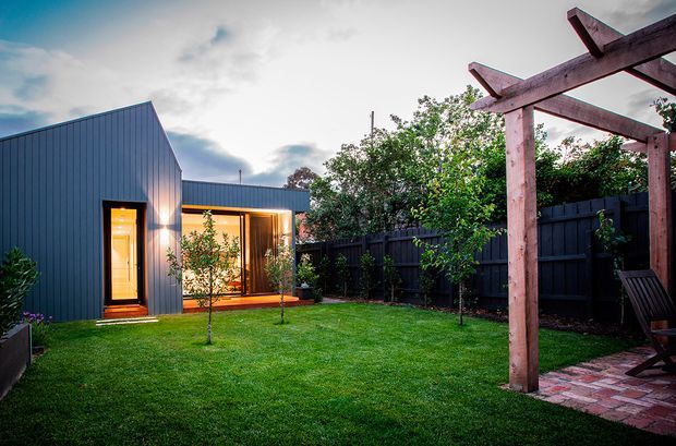Exterior view of a compact contemporary backyard dwelling or extension with dark vertical metal cladding, a steep mono-pitched roofline, and warm illuminated glazing opening onto a manicured lawn. A timber pergola with exposed beams frames a brick-paved sitting area, while dark fencing and mature planting enclose the garden.