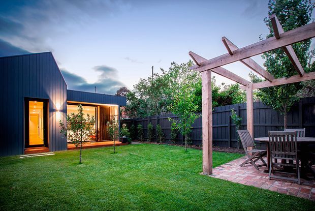 A landscaped backyard viewed from the lawn, featuring a dark-clad modern single-storey dwelling with warm illuminated glazing and a timber pergola over a brick-paved outdoor dining area. The enclosed garden includes lawn, young trees, shrubs, and a dark privacy fence.
