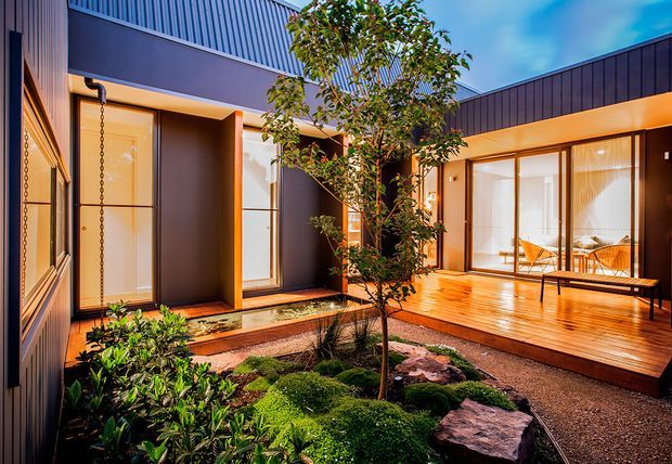 A contemporary courtyard viewed from outside, with dark vertical cladding, warm timber decking, tall glazed doors and windows, and a landscaped central garden featuring a small tree, mossy planting, shrubs, and natural rocks. Warm interior lighting creates an inviting evening atmosphere.