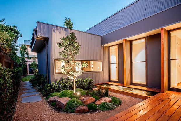 Contemporary single-storey home exterior viewed from a landscaped courtyard. The facade combines dark vertical metal cladding with warm timber-framed floor-to-ceiling glazing and a projecting corrugated metal roof. A timber deck, gravel paths, planted garden beds, and a small rock garden surround the entrance and windows.