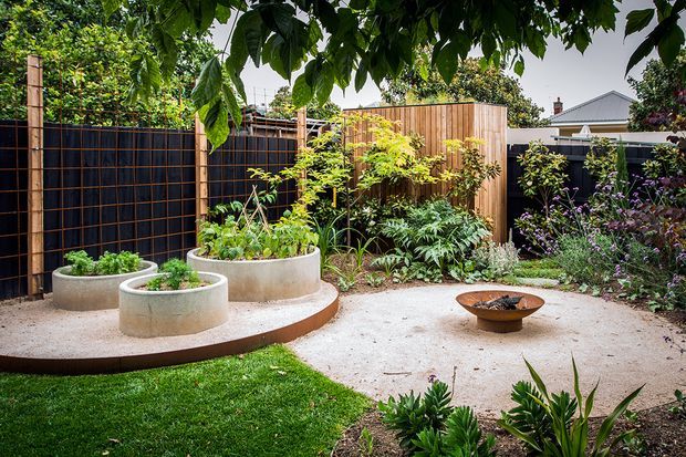 A landscaped residential courtyard garden with curved raised concrete planting beds, lush greenery, timber and dark screened fencing, gravel paving, and a small circular fire bowl. The space has a private, tranquil garden atmosphere with established planting and a suburban home visible beyond the fence.