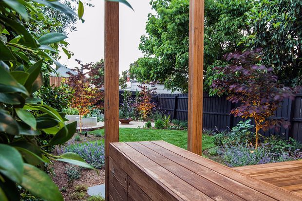 A landscaped residential garden viewed from a timber deck or outdoor entertaining area. The foreground features built-in timber decking and a timber bench or counter beneath a simple timber frame, with layered planting, lawn, raised garden beds and a dark privacy fence beyond.
