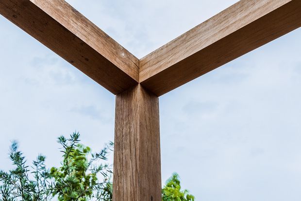 Close-up view of a substantial timber post-and-beam corner structure outdoors, with exposed wood grain and beams forming an open frame against a pale sky and surrounding greenery.