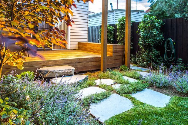 A landscaped residential courtyard garden featuring a raised timber deck with built-in bench seating, timber privacy screening, stepping stones set through low groundcover planting, and dense ornamental shrubs. The space has a warm, natural outdoor feel with timber and pale stone contrasting against lush greenery.
