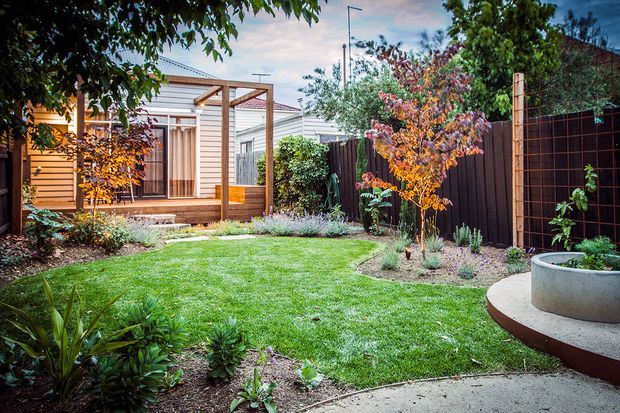 A landscaped residential backyard with a manicured lawn, planted garden beds, mature trees, and a small timber-clad pavilion or outdoor room. The space includes a timber pergola with translucent roofing, timber decking, privacy fencing, curved concrete paving, and a raised circular concrete planter or seating feature.