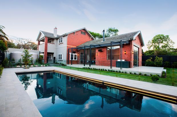 A landscaped residential backyard featuring a large rectangular swimming pool, pale paved pool surround, and a modern glazed outdoor living pavilion. Behind it is a two-storey red-brick and light-rendered house with pitched roofs, dark window frames, and a covered terrace overlooking the pool.