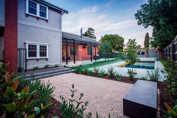 A landscaped courtyard beside a two-storey brick and rendered residential building, featuring a paved garden path, planted borders, a glazed outdoor entertaining area, and a rectangular swimming pool enclosed by glass fencing.