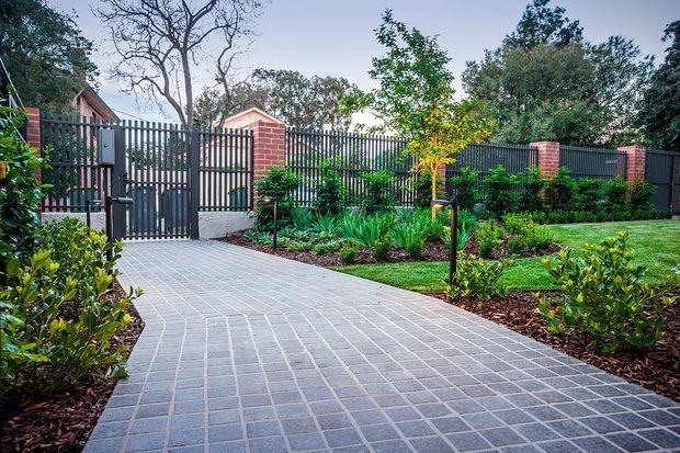 A landscaped residential garden entrance with a broad grey tiled or paved walkway leading to a dark metal pedestrian gate and fence. Brick columns punctuate the boundary, while mulched planting beds, shrubs, ornamental grasses, and a neatly maintained lawn create a structured outdoor setting.