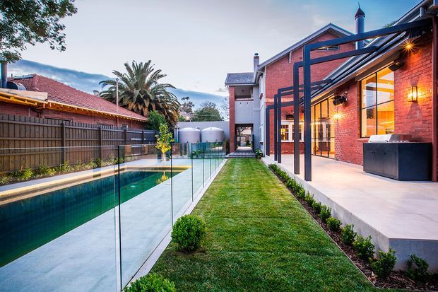 A landscaped residential courtyard viewed from outdoors, featuring a long narrow swimming pool with frameless glass fencing, a manicured lawn, and a contemporary covered entertaining terrace attached to a red-brick house. The terrace has exposed dark steel framing, warm exterior lighting, pale concrete paving, and large glazed openings. A pitched-roof brick dwelling and neighboring fencing frame the enclosed outdoor space.