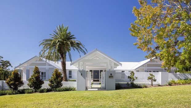 A single-storey white residential building viewed from the front, with multiple pitched gabled roof sections, a central covered entry gate, white picket-style fencing, landscaped shrubs, lawn, and mature palm and deciduous trees under a clear blue sky.