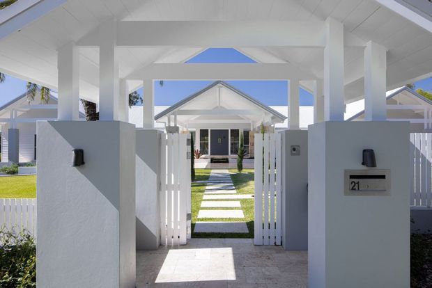 Bright exterior view through a white rendered entry gateway with a covered flat-roof canopy, square posts, white slatted pedestrian gates, and a paved threshold. A straight stepping-stone path crosses a manicured lawn toward a light-coloured gabled house with prominent verandah-like columns and tropical planting. The composition has a clean, symmetrical coastal-residential character.
