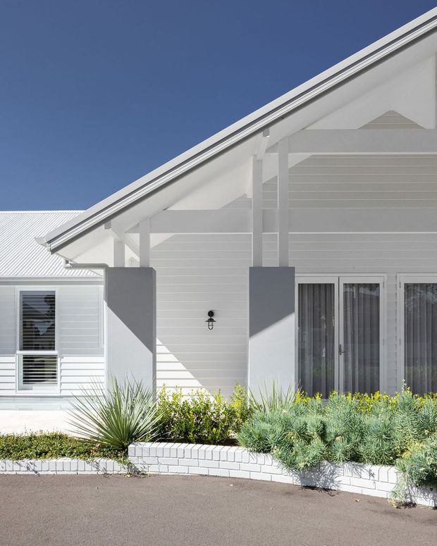 Bright exterior view of a single-storey white weatherboard home with a prominent gabled roof and covered entry or veranda. The facade features white horizontal cladding, white-framed glazing and doors, grey structural columns, and low, drought-tolerant landscaping in raised white brick garden beds.