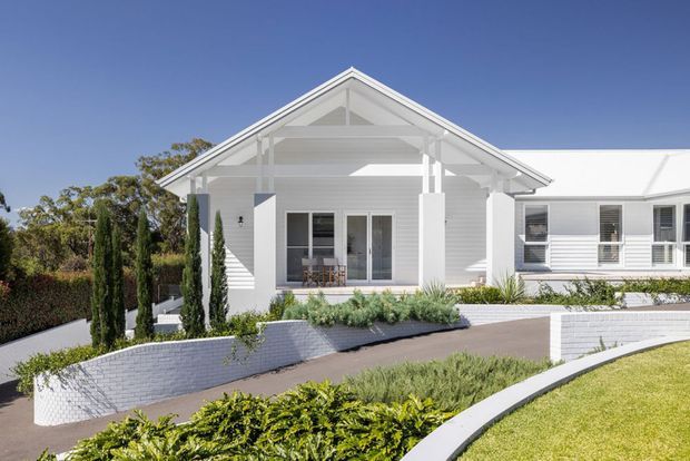 Bright exterior view of a single-storey white weatherboard house with a prominent front-facing gabled roof, covered entry porch, white-framed windows, and landscaped garden beds. Curved rendered or painted masonry retaining walls and a paved driveway/path lead to the entrance.