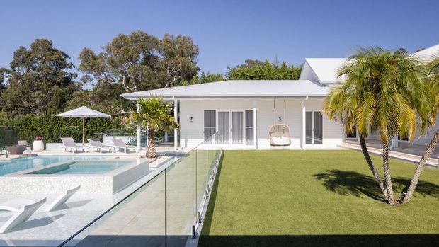 A bright single-storey white residence with a low pitched roof, extensive glass doors, manicured lawn, palm trees, and a tiled swimming pool enclosed by frameless glass balustrades. Poolside loungers and umbrellas create a resort-like outdoor setting.