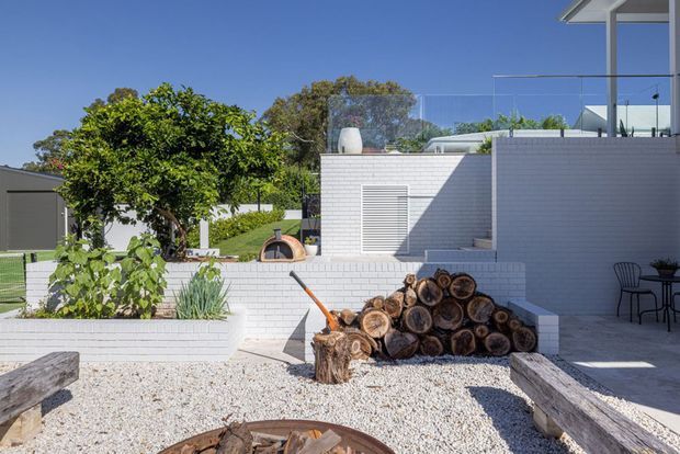 Bright contemporary residential courtyard with white painted brick walls, gravel landscaping, raised garden beds, stacked firewood, timber bench seating, and a small outdoor pizza oven. A modern multi-level home with glass balustrades and a rooftop terrace is visible behind the courtyard.