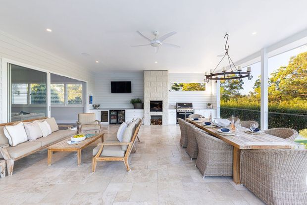 Bright, airy enclosed outdoor entertaining room with white horizontal wall panelling, large glazed openings, a central stone fireplace, and an open kitchen/barbecue area. The space combines a relaxed lounge with a wicker dining setting, light timber furniture, pale stone flooring, ceiling fan, and a rustic chandelier. Views of greenery are visible through the glazing.