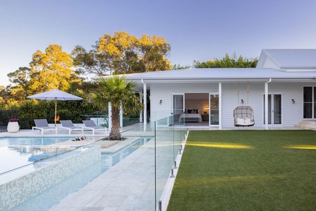 A bright single-storey white-clad residence viewed from the pool area, with a long covered veranda, glass pool fencing, pale tiled pool deck, manicured lawn, outdoor lounge chairs, umbrella, palm planting, and a hanging egg chair. The home has a low corrugated roof and expansive glazed openings, with a living room visible through the central opening.