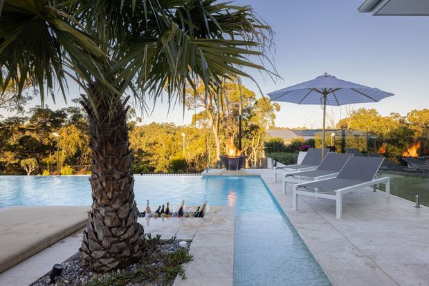 Luxury outdoor pool area with a narrow tiled water channel, pale stone paving, white sun loungers, a large grey umbrella, fire features, and a prominent palm tree overlooking a landscaped setting.
