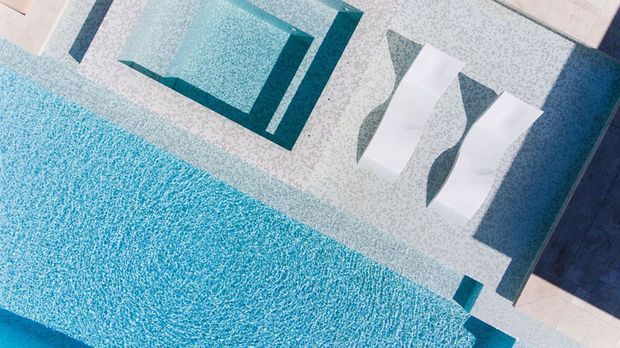 A top-down view of a contemporary swimming pool with vivid turquoise water, geometric inset sections, pale patterned paving, and two white in-water loungers. The design has crisp angular lines and a cool blue-and-white palette.