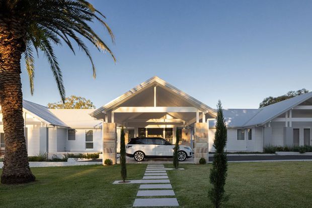 Front exterior view of a low-rise residential property with a prominent central gabled porte-cochère and covered driveway, flanked by white weatherboard wings with matching pitched roofs. A neat lawn, clipped ornamental trees, stepping-stone path, and palm tree frame the symmetrical facade.