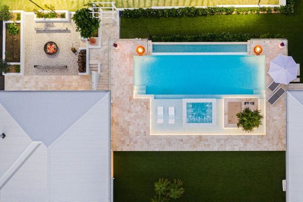 Aerial top-down view of a landscaped residential courtyard with a rectangular swimming pool, paved stone terrace, sun loungers, parasols, a small spa or water feature, and an outdoor fire pit seating area. The courtyard is framed by light-coloured tiled roofs, manicured lawns, hedges, and garden planting.