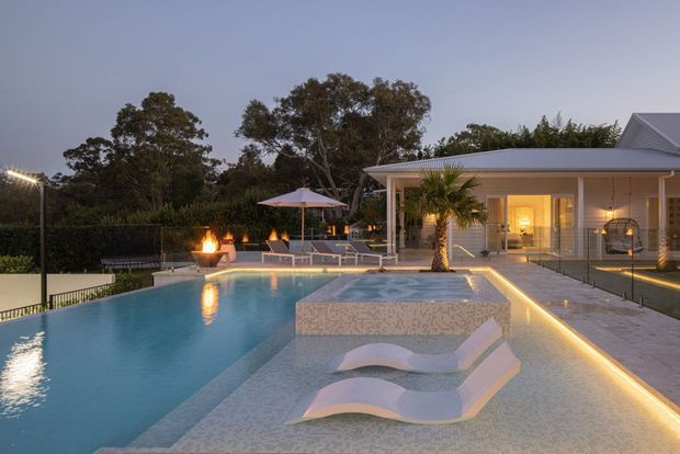 Twilight view of a landscaped residential pool area with a large swimming pool, shallow sun shelf, curved in-water loungers, illuminated water feature, outdoor seating, fire bowls, and a single-storey light-coloured pavilion with a covered terrace and pitched roof.