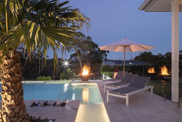 Twilight view of a refined outdoor pool terrace with a rectangular swimming pool, illuminated shallow ledge, sculptural fire bowls, sun loungers, a large umbrella, tropical palms and landscaped surroundings. The terrace is adjacent to a modern building with pale rendered walls and square columns.