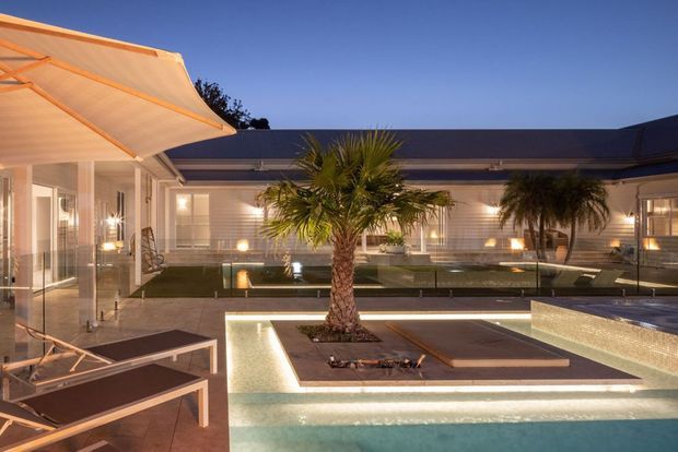 Dusk view of a refined courtyard pool area framed by a low, dark-roofed building. The scene includes a large illuminated swimming pool, shallow water features, a central palm planting bed, glass balustrades, paved terraces, and a cantilevered outdoor canopy with warm integrated lighting.