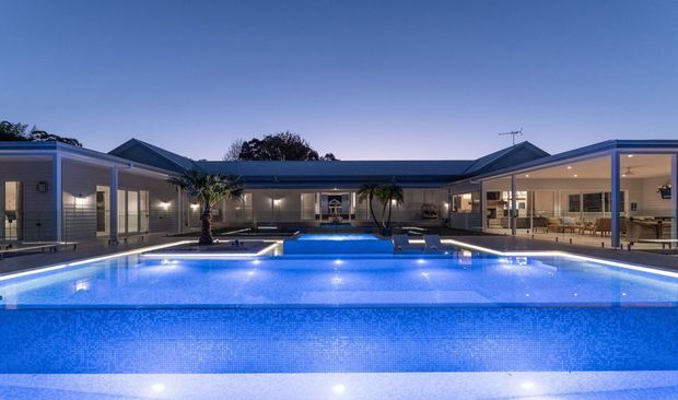 Dusk exterior view of a low, single-storey contemporary residence arranged around a large illuminated swimming pool. The home has pale horizontal cladding, dark pitched roof sections, extensive floor-to-ceiling glazing, and an expansive open-plan living and dining area visible on the right. Poolside palms and integrated lighting create a resort-like atmosphere.