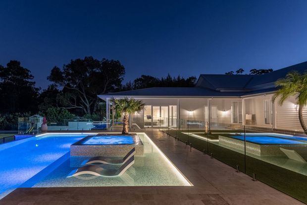 Dusk exterior view of a contemporary single-storey residence with a large illuminated swimming pool, raised spa feature, shallow water element, glass pool fencing, paved terrace, palm trees and a low pitched roof with light horizontal cladding.